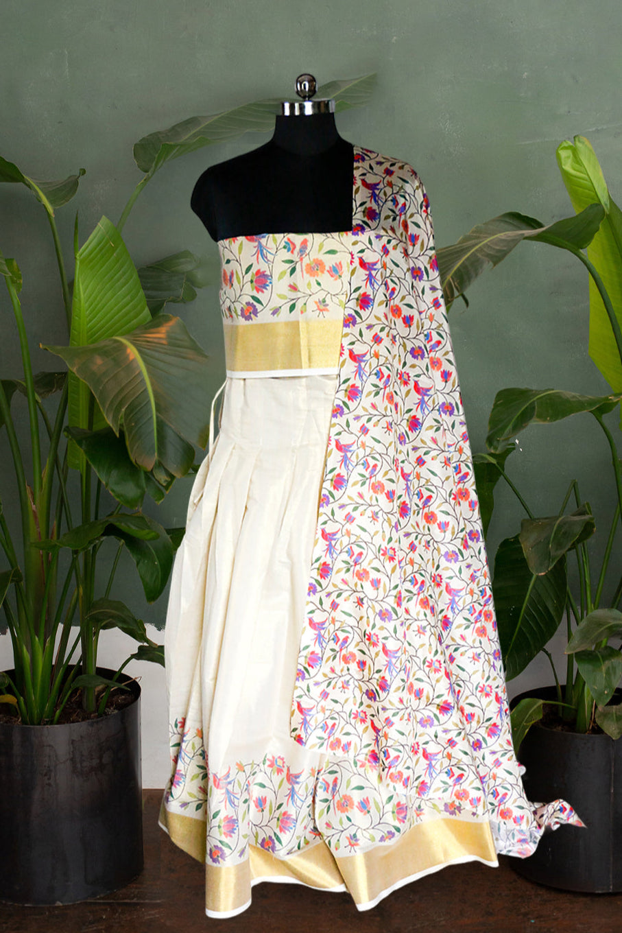 Mannequin wearing a floral dress with a patterned shawl against a green wall with plants.
