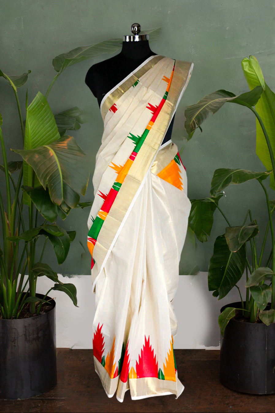 White saree with colorful patterns on a mannequin against a green wall with plants.