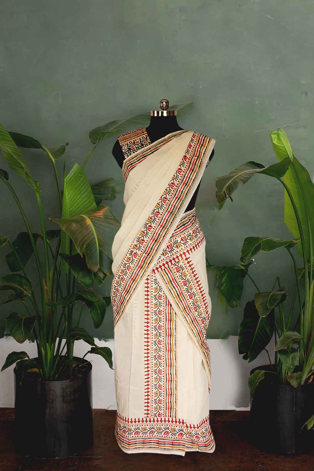 Saree with intricate patterns on a mannequin against a green wall with plants.