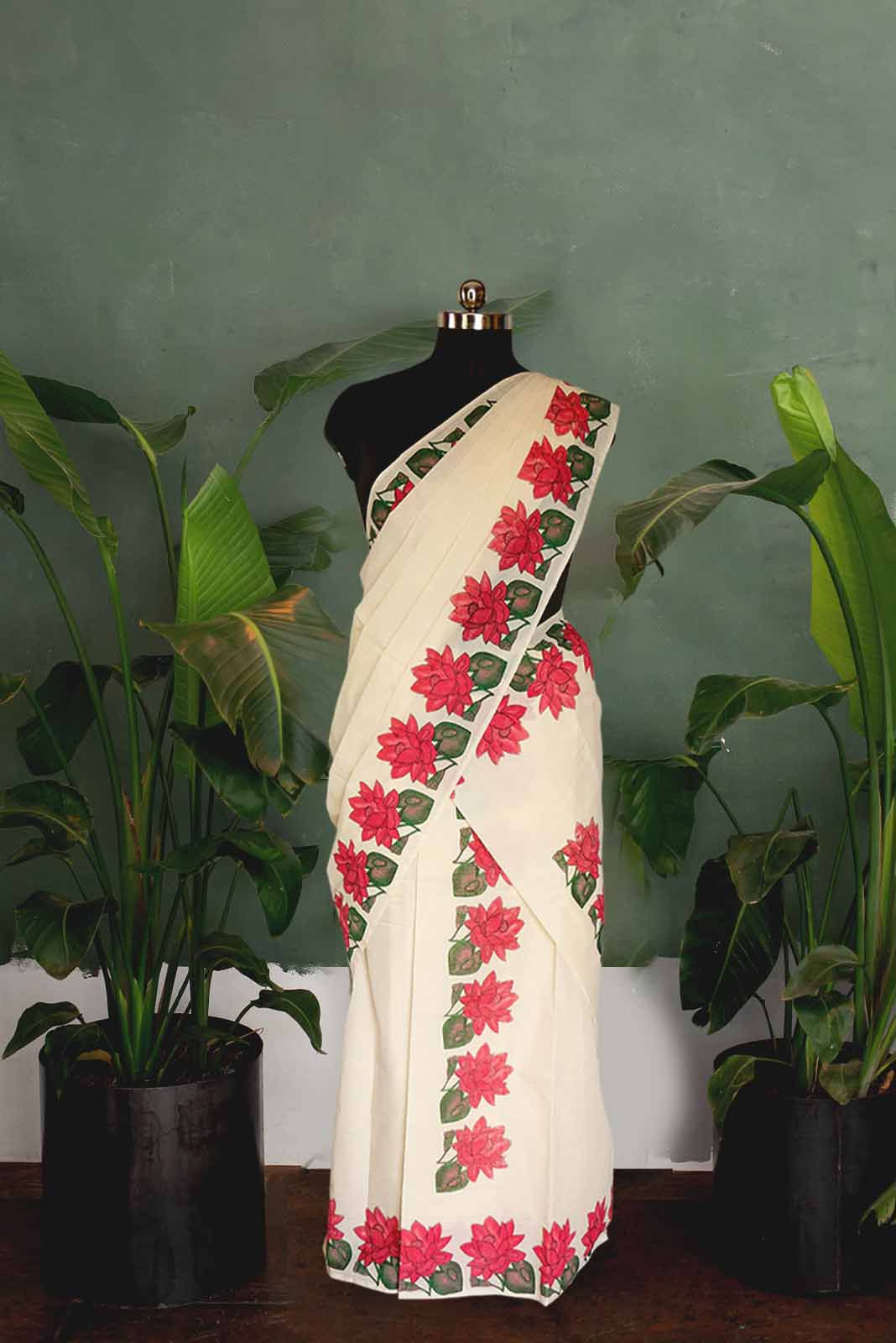 White saree with red floral patterns on a mannequin against a green wall with plants.